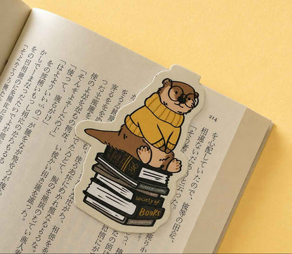 A die-cut sticker featuring an illustration of an otter wearing round glasses and a yellow sweater, sitting on a stack of books, placed on an open book with Japanese text against a warm yellow background.