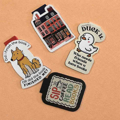 Four colorful book-themed stickers arranged on a solid tan background. The stickers feature illustrations of a dog, a library building, a white duck, and a coffee shop sign with text like 'Duo k it', 'Who needs a friend when you have books', and 'SIP COFFEE READ BOOKS'.