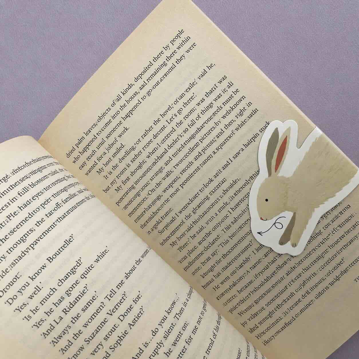 A die-cut paper bookmark shaped like a beige bunny with pink inner ears and a black nose, placed on an open book page against a lavender background.