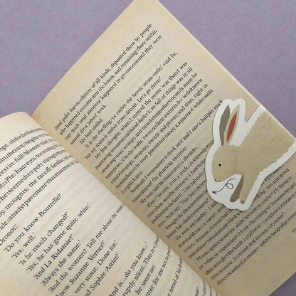 A die-cut paper bookmark shaped like a beige bunny with pink inner ears and a black nose, placed on an open book page against a lavender background.