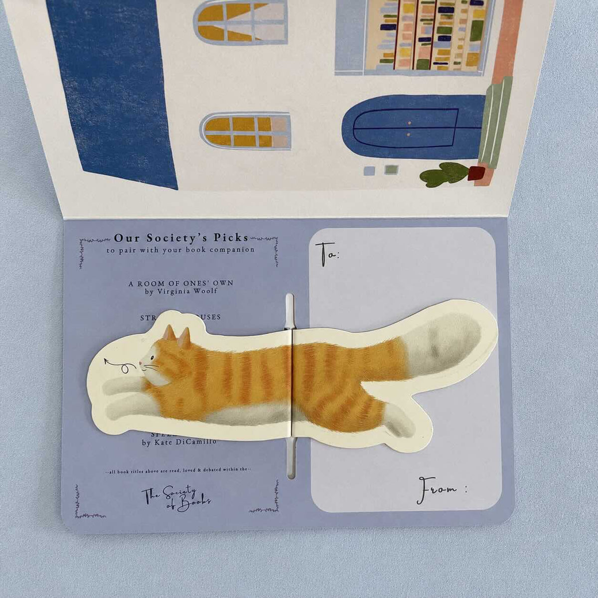 An open illustrated book or card featuring a die-cut orange tabby cat illustration across the center, with printed text and address fields on the pages. The background is a soft light blue surface.