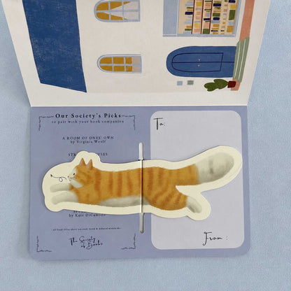 An open illustrated book or card featuring a die-cut orange tabby cat illustration across the center, with printed text and address fields on the pages. The background is a soft light blue surface.