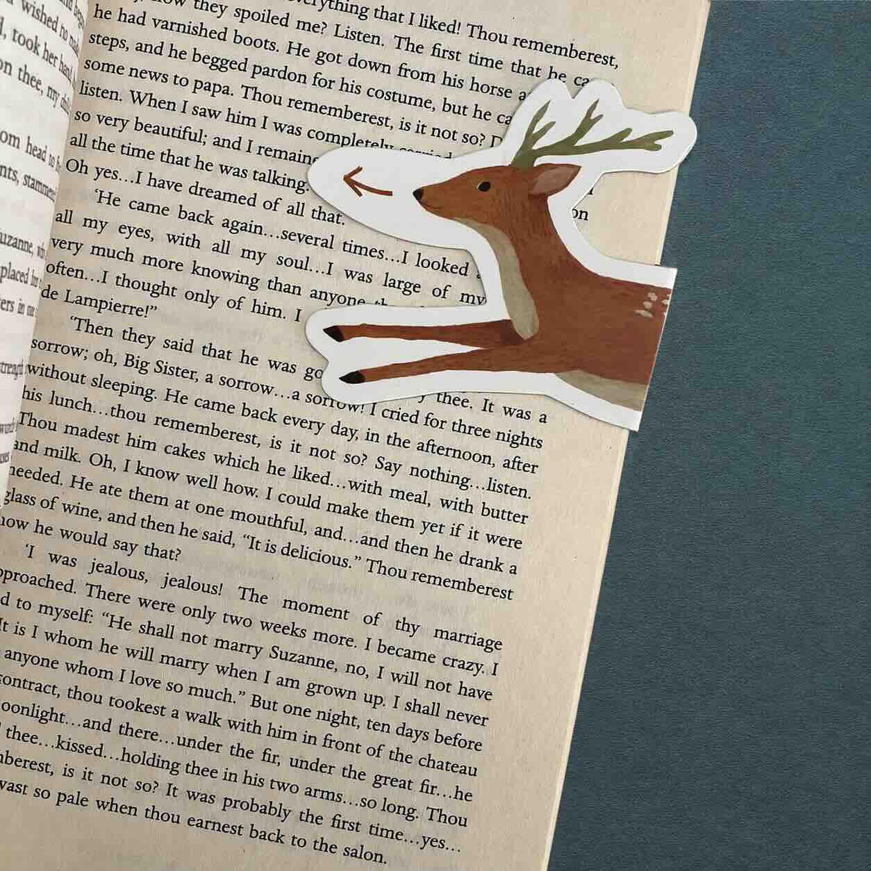 A handmade paper cutout of a reindeer with antlers, placed over an open book page. The reindeer has a light brown body, a dark brown face and ears, and a white antlered head.
