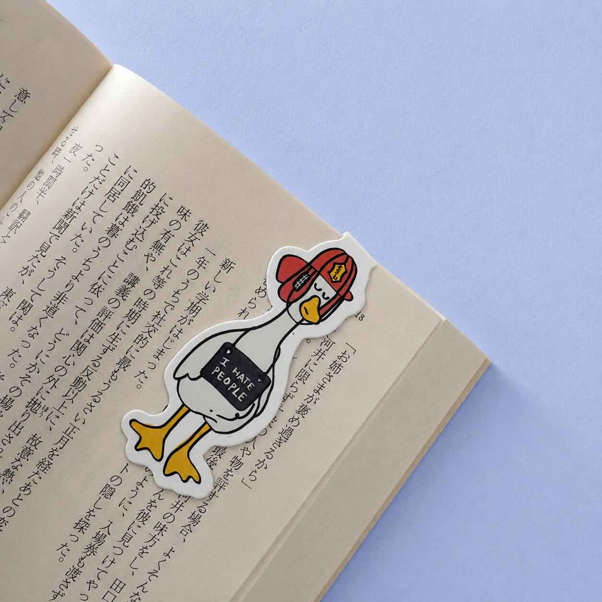 A cartoon duck sticker with a red beret and a black sign reading 'I HATE PEOPLE' lies on an open book with printed text, set against a light blue background.