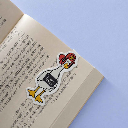 A cartoon duck sticker with a red beret and a black sign reading 'I HATE PEOPLE' lies on an open book with printed text, set against a light blue background.