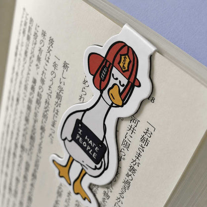 A die-cut paper bookmark shaped like a cartoon duck wearing a red cap and holding a sign that says 'I HATE PEOPLE,' clipped onto an open book with Japanese text.