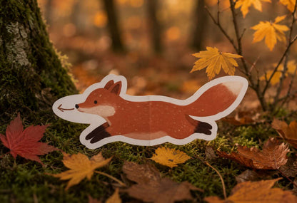 A die-cut fox sticker or sign with a white border, featuring an orange-brown fox with black-tipped paws and tail tip, placed on mossy ground with autumn leaves and a tree trunk in a forest setting.