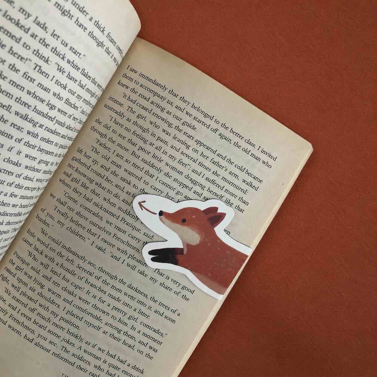 A die-cut sticker of a cartoon fox bookmark is placed on an open book page, set against a solid reddish-brown background.