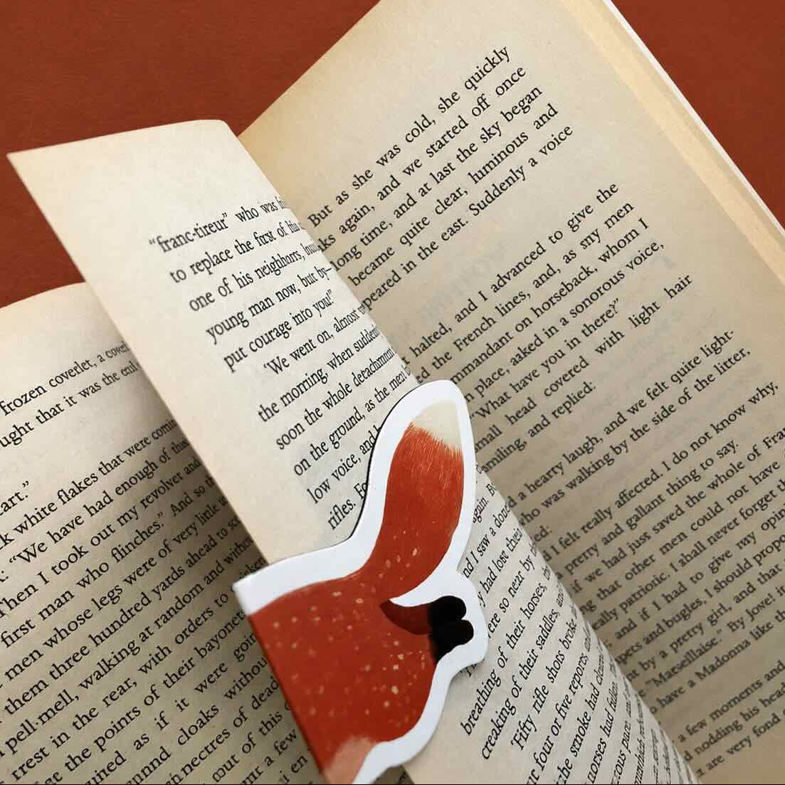 A small fox-shaped bookmark with a red-orange body, white outline, and black nose rests on open book pages against a warm brown background.