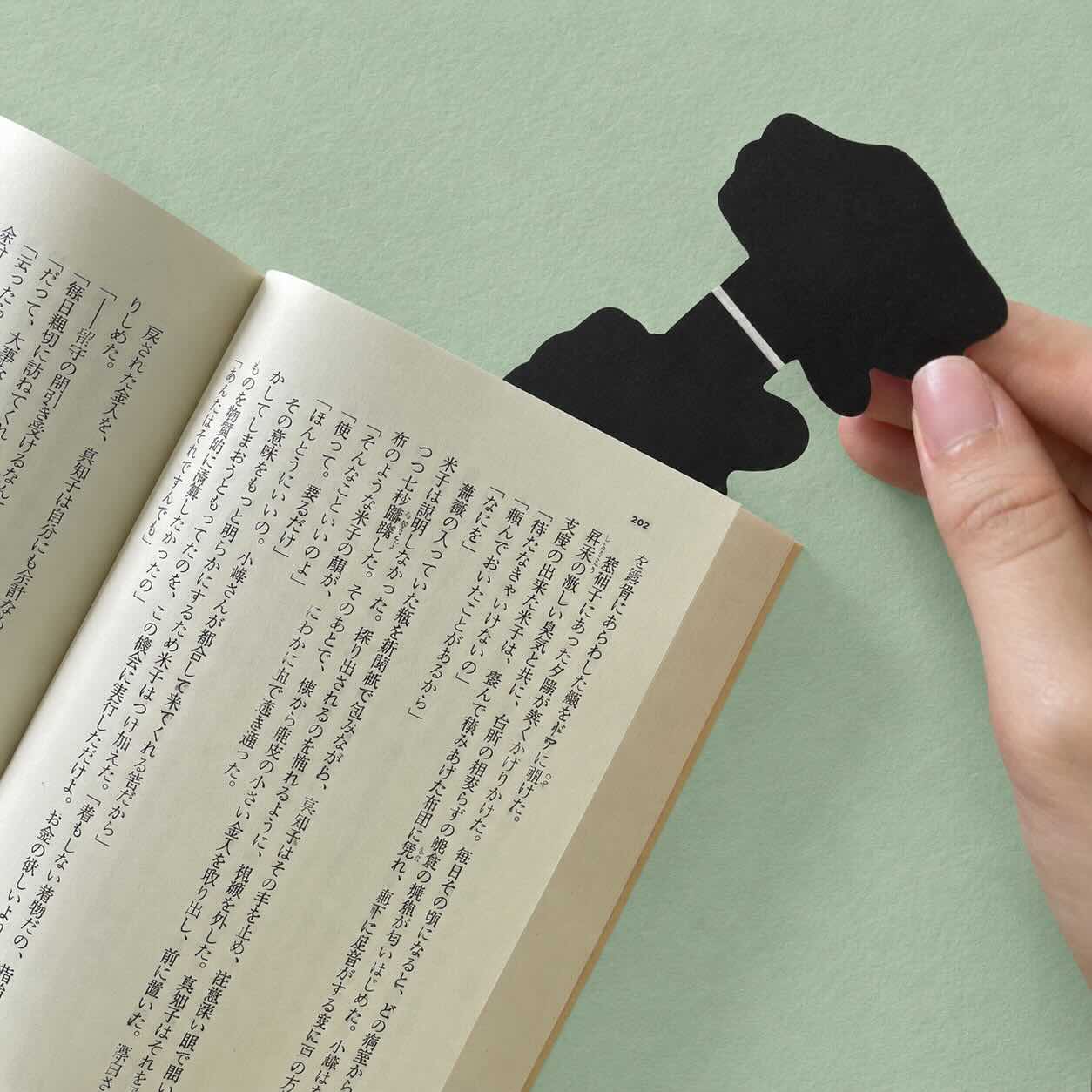 A hand holds a black bookmark shaped like a hand against the pages of an open book on a light green background.