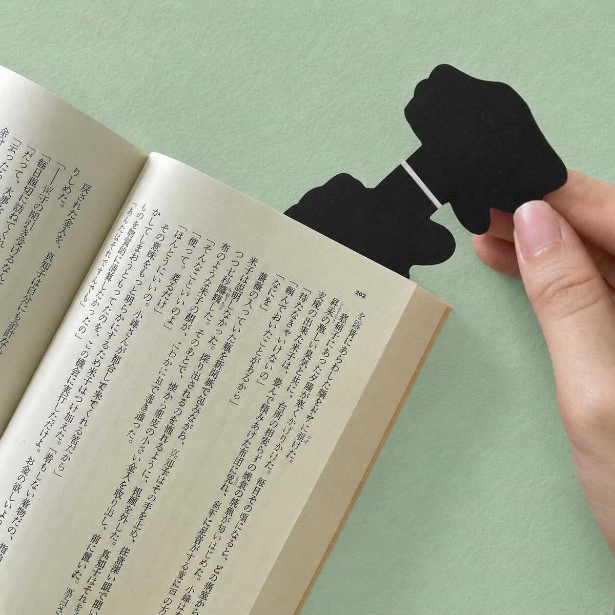 A hand holds a black bookmark shaped like a hand against the pages of an open book on a light green background.