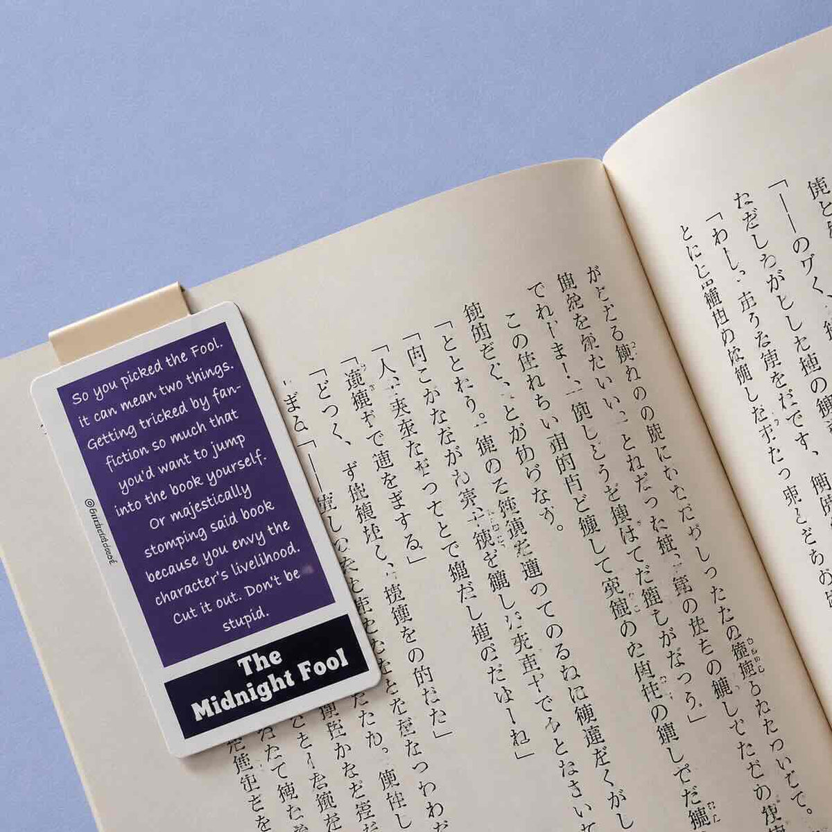 A small rectangular card or bookmark with a purple front and black text reading 'The Midnight Fool' is placed on an open book with printed text in Japanese. The card is photographed on a pale blue background.