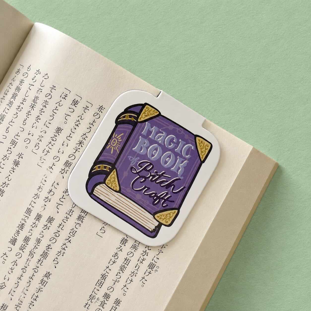 A die-cut sticker shaped like a purple magic book with gold corner accents and the words 'Magic Book Bitch Craft' printed on it, placed on an open book with Japanese text against a light green background.