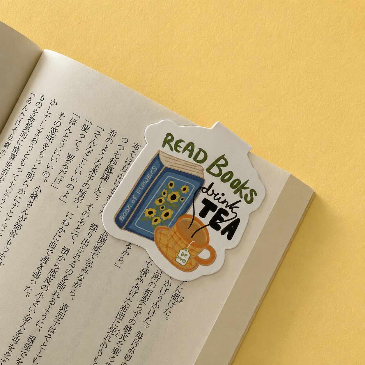 A die-cut sticker with the text 'READ BOOKS drink TEA' and an illustration of a book, sunflowers, and a teacup, placed on an open book page against a yellow background.