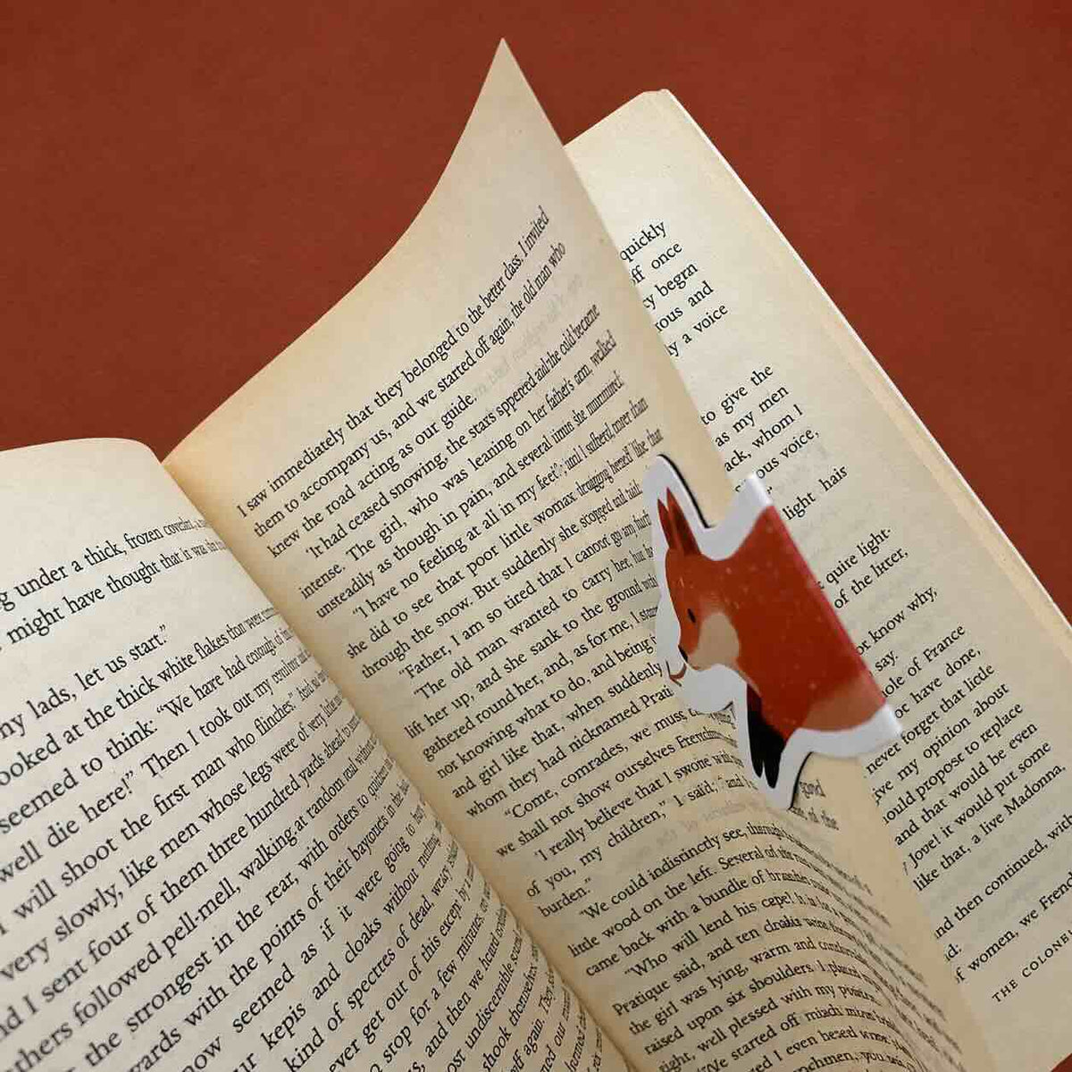 A small fox-shaped bookmark made from a book page, featuring a red-orange fox head and body with black and white details, shown inside an open book on a reddish-brown background.