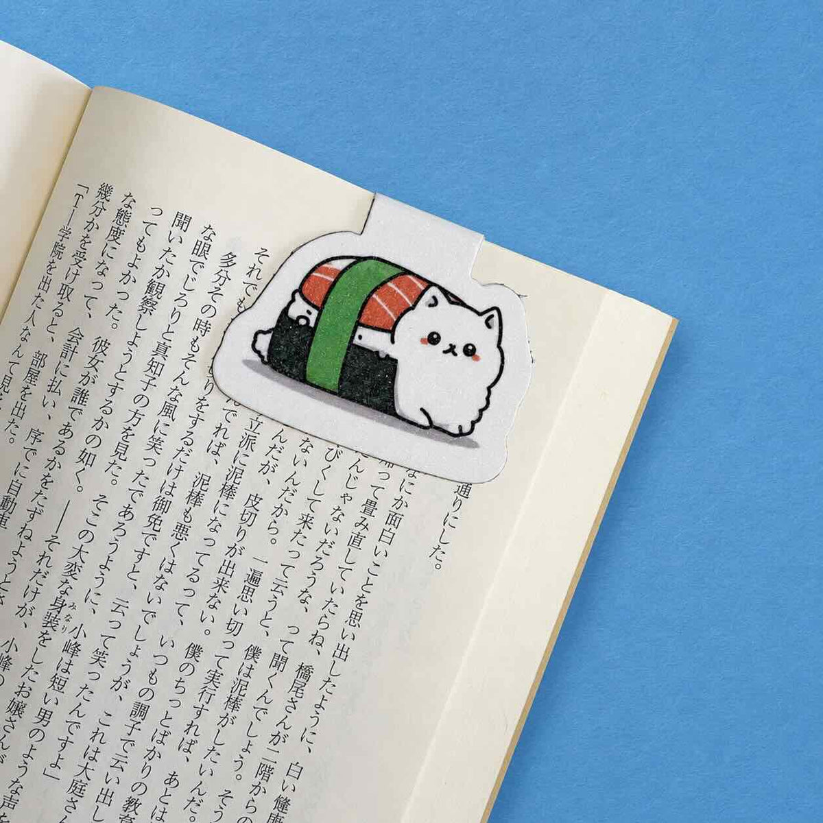 A die-cut sticker magnetic bookmark featuring a cute white cat dressed as sushi, placed on an open book with Japanese text against a solid blue background.