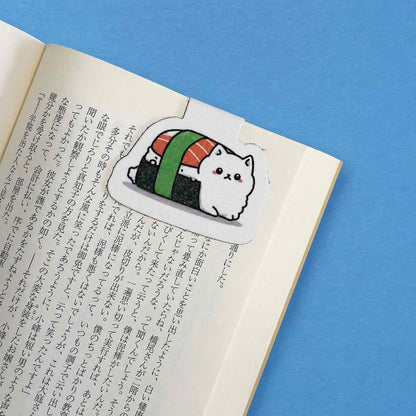 A die-cut sticker magnetic bookmark featuring a cute white cat dressed as sushi, placed on an open book with Japanese text against a solid blue background.